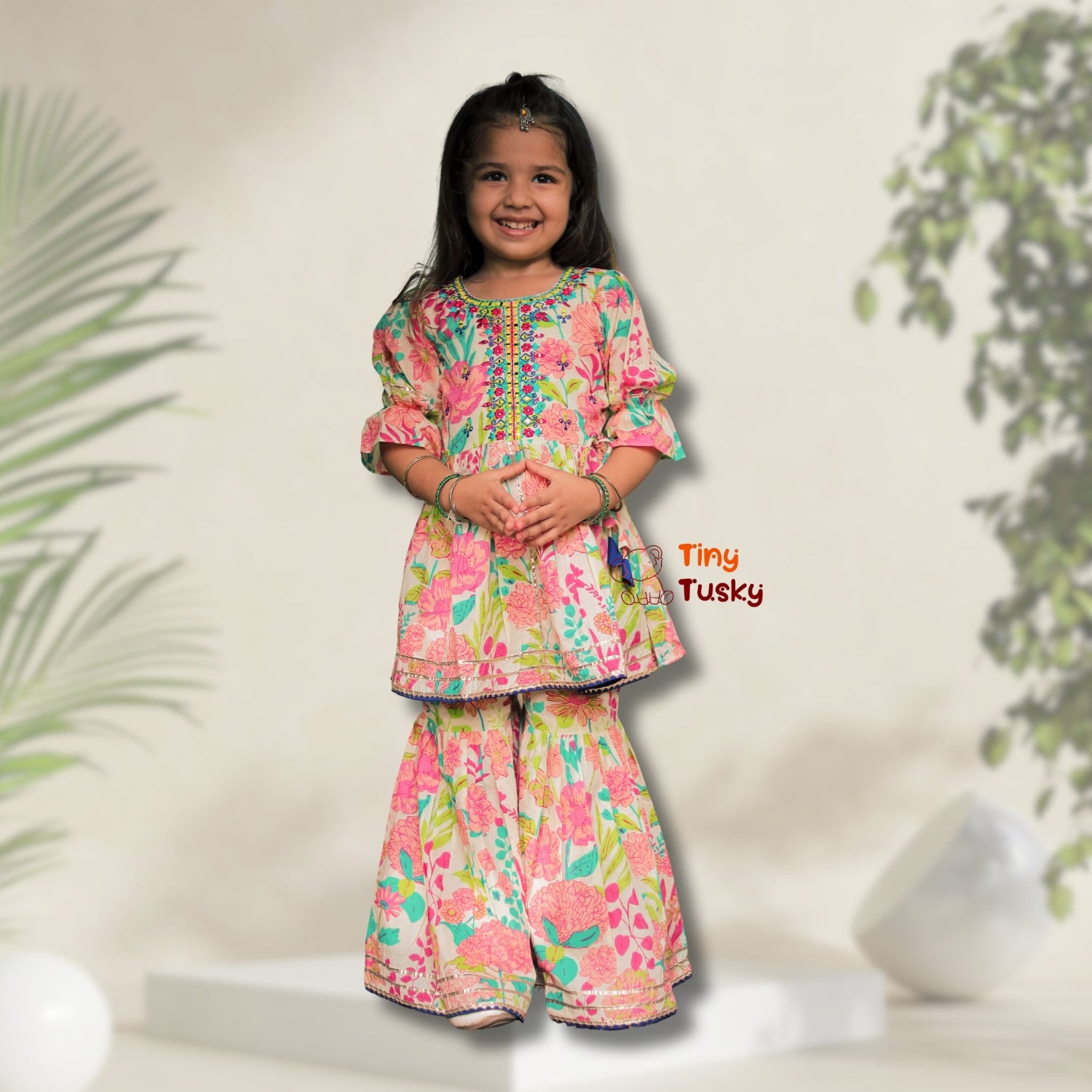 Young girl wearing a colorful floral outfit with Tiny Tusky branding, standing against a neutral background with plants.